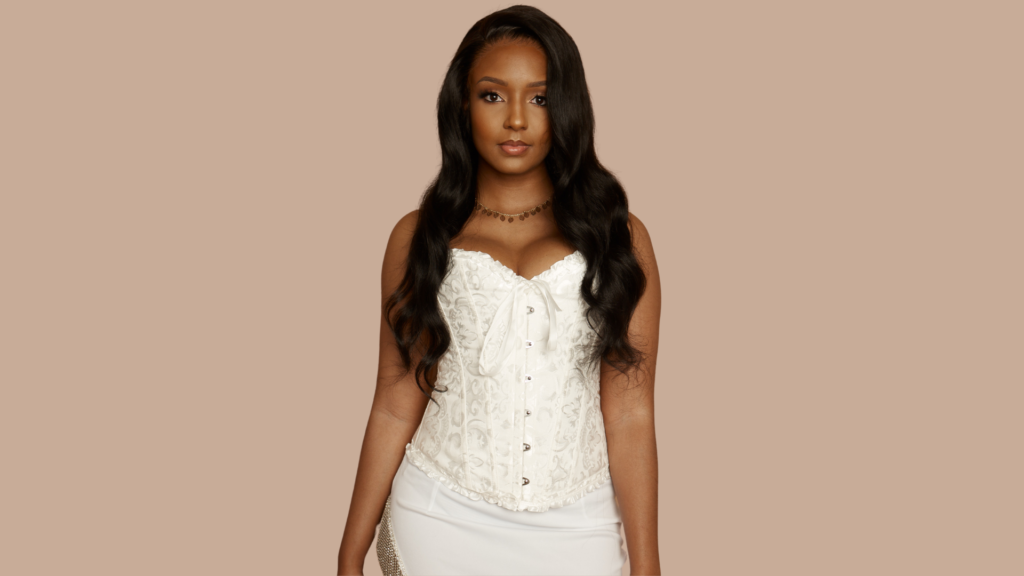 Woman in a white corset top with long, wavy hair, showcasing a confident look, representing custom wigs and bridal styles at Indish Beauty.