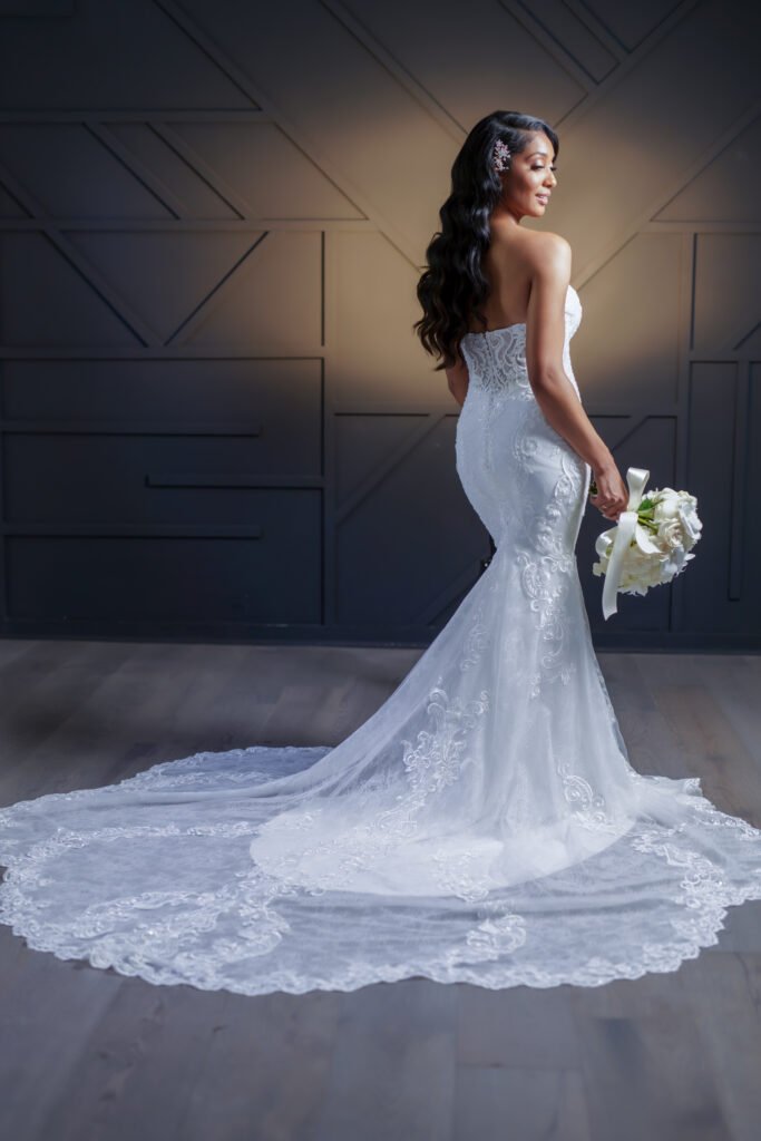 Bridal model in elegant lace gown showcasing long, flowing hair with floral accessory, holding bouquet, against stylish backdrop, emphasizing wedding hair and beauty services at Indish Beauty.